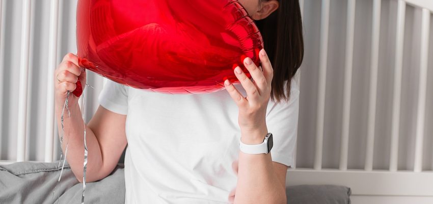 Woman sitting on bed with red baloon in shape of heart and gift box with bouquet gypsophila.